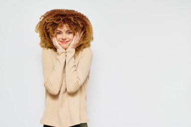 Bej gömlekli, kıvırcık saçlı mutlu genç bir kadın neşeli bir tavır sergiliyor. Minimalist beyaz bir arkaplanda duruyor, neşeli ekspres yoluyla neşe ve pozitiflik hissini iletiyor.