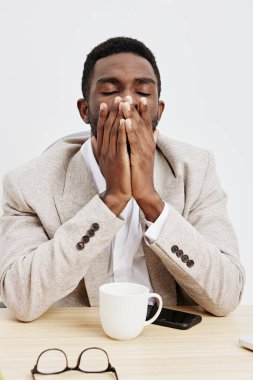 Yorgun ve stresli genç bir adam koyu tenli, gözleri kapalı, burnu havada, kahve fincanı, akıllı telefon ve gözlüklerle masada oturuyor, yorgunluğu, tükenmişliği, tükenmişliği, günlük rutini,