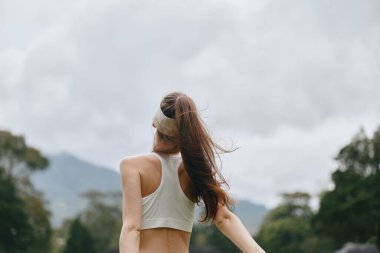 Kafkas kökenli, sportif bir elbise giyen sağlıklı bir kadın kollarını açık havada neşeyle uzatıyor. Doğal arka plan ve bulutlu gökyüzü sağlık ve canlılığı vurguluyor.