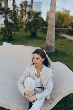 Açık renk elbiseli genç bir kadın bahar parkındaki eğri taşlı bankta düşünceli bir şekilde oturuyor. Doğal yeşil arka planlı moda ve psikolojik konsepti çağrıştırıyor..