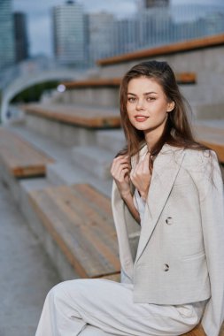 Açık renk ceket ve beyaz pantolon giyen şık genç bir kadın şehir merdivenlerinde oturuyor, kendine güvenini ve zarafetini doğal makyajıyla ve gün ışığıyla, moda tarzıyla gösteriyor.