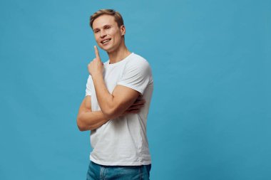 Sarı saçlı, sıradan beyaz tişörtlü, mavi arka planda duran mutlu genç adam, bir jest yapıyor, parmağını yukarı kaldırıyor, pozitifliği iletiyor, düşünüyor ya da bir çözüm öneriyor.