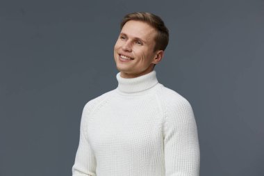 Beyaz balıkçı yaka kazak giymiş gülümseyen bir adamın portresi, düz gri bir zemin üzerinde, neşeli ve rahat bir tavır sergiliyor, moda, yaşam tarzı ve portre konseptleri için uygun.