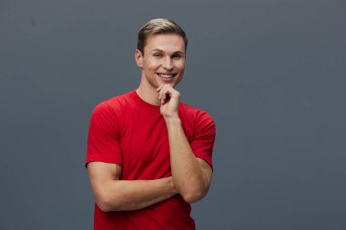 Kırmızı t-shirt giyen gülümseyen genç bir adamın portresi, kolları çapraz ve çenesinde duran, mutluluk ve kendine güveni ifade eden, sade gri arka plan, gündelik tarz, neşeli ruh hali.