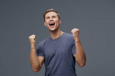 Mutlu adam kendini muzaffer ve enerjik hissediyor, günlük kıyafet, coşkuyla gülümsüyor, başarıyı kutluyor, sade gri fon, stüdyo ortamı, pozitif ifade. Kavram: Başarı ve