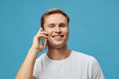 Arkadaşça gülümseyen bir adam bir telefon görüşmesi yapıyor, beyaz bir tişört giyiyor, parlak mavi bir arka plana karşı duruyor, günlük tarz, pozitif ruh hali, iletişim, modern yaşam tarzı, mutluluk, teknoloji