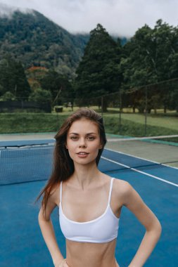Beyaz spor sutyenli enerjik genç bir kadın yemyeşil bir alanla çevrili bir tenis kortuna güvenerek sağlık ve atletizmi vurguluyor.