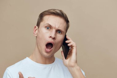 Şok olmuş genç bir adam telefon kulaklığı ile konuşuyor, ağzı açık ve ifade edici bir yüz ifadesiyle bej arka plan yaşam tarzı konseptine karşı günlük beyaz tişört giyiyor dijital detoks ve aletlerle akıl sağlığı.