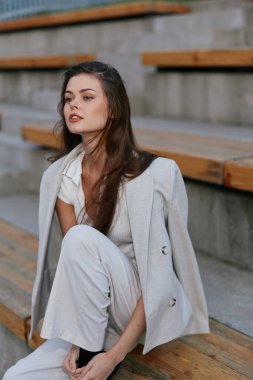 Açık renk takım elbiseli genç bir kadının portresi, tahta merdivenlerde oturuyor, doğal bir yüz ifadesi, doğal bir makyaj ve rahat bir poz sergiliyor..