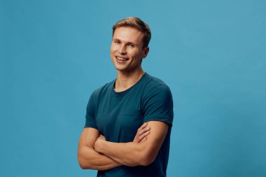 Mavi tişörtlü gülümseyen beyaz adam kollarını kavuşturmuş mavi arka plandaki kameraya bakıyor. Siber güvenlik ve dijital korumayı temsil eden kendinden emin mutlu erkek portresi