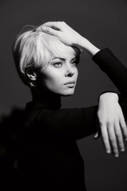 Zarif kısa sarı saçlı, zarif genç bir kadın koyu bir arka planda dramatik bir şekilde poz veriyor, kendine güveni ve duygusal derinliği gösteriyor..