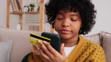 African american woman shopping online holding smartphone paying with gold credit card. Girl sitting at home buying on Internet enter credit card details. Online shopping ecommerce delivery service