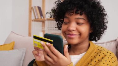 African american woman shopping online holding smartphone paying with gold credit card. Girl sitting at home buying on Internet enter credit card details. Online shopping ecommerce delivery service