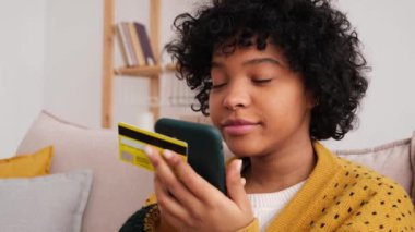 African american woman shopping online holding smartphone paying with gold credit card. Girl sitting at home buying on Internet enter credit card details. Online shopping ecommerce delivery service