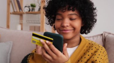 African american woman shopping online holding smartphone paying with gold credit card. Girl sitting at home buying on Internet enter credit card details. Online shopping ecommerce delivery service