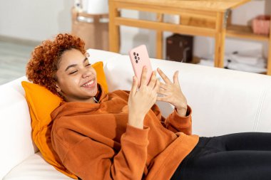 African american girl lying on couch with smartphone having video chat. Female blogger chatting with best friends in social network. Young woman having virtual meeting online chat video call at home