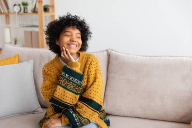 Beautiful african american girl with afro hairstyle smiling sitting on sofa at home indoor. Young african woman with curly hair laughing. Freedom happiness carefree happy people concept