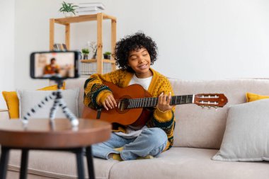Blogger guitarist. African american girl blogger playing guitar talking to webcam recording vlog. Social media influencer woman streaming at home indoors. Music content creator broadcast tutorial