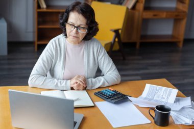 Middle aged senior woman sit with laptop and paper document. Pensive older mature lady reading paper bill pay online at home managing bank finances calculating taxes planning loan debt pension payment