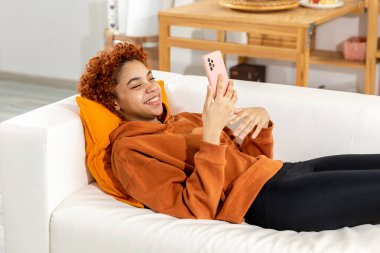 African american girl lying on couch with smartphone having video chat. Female blogger chatting with best friends in social network. Young woman having virtual meeting online chat video call at home