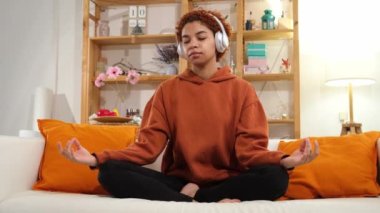 Yoga farkındalığı meditasyonu. Genç sağlıklı Afrikalı kız evde yoga yapıyor. Lotus 'ta oturan bir kadın kanepeye oturmuş, meditasyon yapıyor, gülümsüyor, evde dinleniyor. Nefes egzersizi yapan bir kız. Evde yoga.
