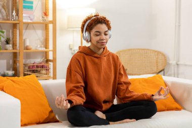 Yoga mindfulness meditation. Young healthy african girl practicing yoga at home. Woman sitting in lotus pose on couch meditating smiling relaxing indoor. Girl doing breathing practice. Yoga at home
