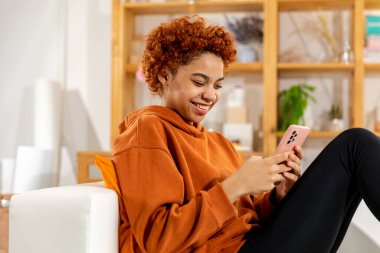 African girl holding smartphone touch screen typing scroll page at home. Woman with cell phone surfing internet using social media apps playing game. Shopping online Internet news cellphone addiction