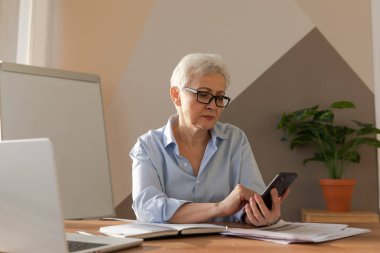 Confident stylish european middle aged senior woman using smartphone at workplace. Stylish older mature 60s gray haired lady businesswoman with cell phone in office. Boss leader using internet apps