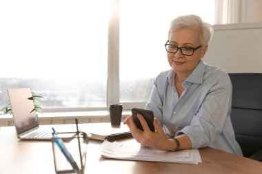 Confident stylish european middle aged senior woman using smartphone at workplace. Stylish older mature 60s gray haired lady businesswoman with cell phone in office. Boss leader using internet apps