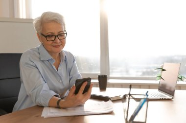 İş yerinde akıllı telefon kullanan kendine güvenen Avrupalı orta yaşlı bir kadın. Tarz sahibi, olgun, 60 'lı yaşlarda, ofisinde cep telefonu olan beyaz saçlı bir iş kadını. Patron internet uygulamaları kullanıyor