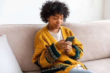 African girl holding smartphone touch screen typing scroll page at home. Woman with cell phone surfing internet using social media apps playing game. Shopping online Internet news cellphone addiction