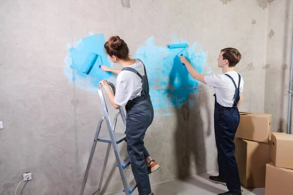 House decoration DIY renovation concept. Happy couple in empty room of new home painting wall together. Husband and wife holding paint roller painting wall with blue color paint in new house