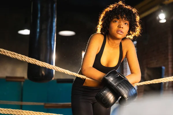 Woman fighter girl power. African american woman fighter with boxing gloves standing on boxing ring leaning on ropes waiting and resting. Strong powerful girl. Strength fit body workout training