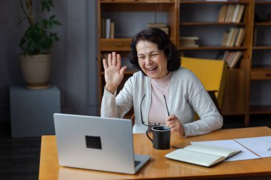 Orta yaşlı mutlu bir kadın dizüstü bilgisayarla oturup arkadaşlarıyla görüntülü konuşma yapıyor. Yaşlı ve olgun büyükanneme gülüp yetişkin çocuklarla internette konuşarak eğleniyorum.