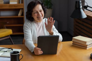 Arkadaşlarıyla video görüşmesinde elinde tablet olan mutlu orta yaşlı bir kadın. Yaşlı ve olgun büyükanneme gülüp yetişkin çocuklarla internette konuşarak eğleniyorum.