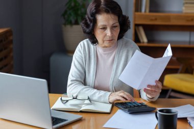Middle aged senior woman sit with laptop and paper document. Pensive older mature lady reading paper bill pay online at home managing bank finances calculating taxes planning loan debt pension payment