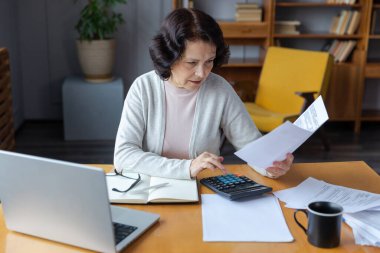 Middle aged senior woman sit with laptop and paper document. Pensive older mature lady reading paper bill pay online at home managing bank finances calculating taxes planning loan debt pension payment