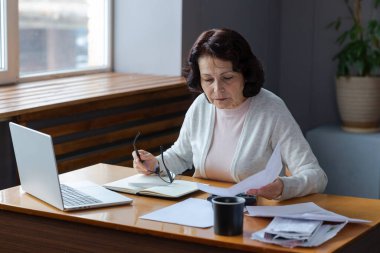 Orta yaşlı yaşlı bir kadın dizüstü bilgisayarı ve kağıt belgeleriyle oturuyor. Düşünceli, olgun, kağıt faturası okuyan, ev idaresi, banka hesapları, kredi emekliliği ödemesi planlayan vergileri finanse eden yaşlı bir kadın.