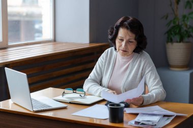 Middle aged senior woman sit with laptop and paper document. Pensive older mature lady reading paper bill pay online at home managing bank finances calculating taxes planning loan debt pension payment