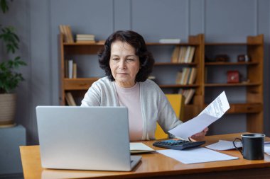 Middle aged senior woman sit with laptop and paper document. Pensive older mature lady reading paper bill pay online at home managing bank finances calculating taxes planning loan debt pension payment