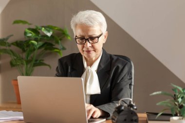 İş yerinde dizüstü bilgisayar kullanan kendine güvenen Avrupalı orta yaşlı bir kadın. Modaya uygun, olgun 60 'ların beyaz saçlı iş kadını ofis masasında oturuyor. Patron öğretmen profesyonel işçi.