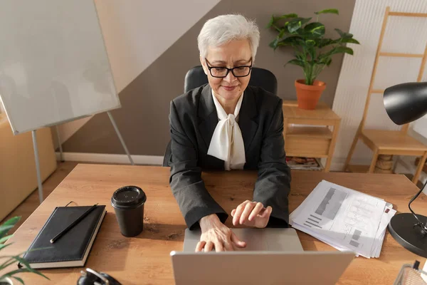 stock image Confident stylish european middle aged senior woman using laptop at workplace. Stylish older mature 60s gray haired lady businesswoman sitting at office table. Boss leader teacher professional worker