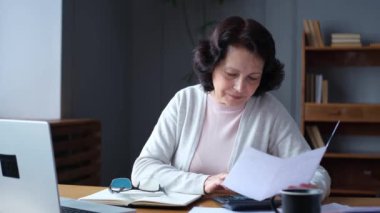 Middle aged senior woman sit with laptop and paper document. Pensive older mature lady reading paper bill pay online at home managing bank finances calculating taxes planning loan debt pension payment