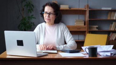 Middle aged senior woman sit with laptop and paper document. Pensive older mature lady reading paper bill pay online at home managing bank finances calculating taxes planning loan debt pension payment