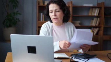 Middle aged senior woman sit with laptop and paper document. Pensive older mature lady reading paper bill pay online at home managing bank finances calculating taxes planning loan debt pension payment