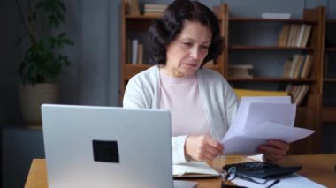 Middle aged senior woman sit with laptop and paper document. Pensive older mature lady reading paper bill pay online at home managing bank finances calculating taxes planning loan debt pension payment