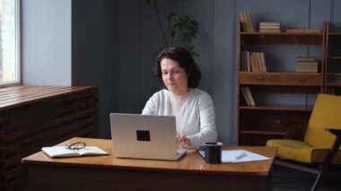 Evde dizüstü bilgisayar kullanan kendine güvenen Avrupalı orta yaşlı bir kadın. Yaşlı, şık 60 'lı yaşlarda bir kadın masada oturmuş bilgisayar ekranına bakıyor daktilo yazarak sohbet ediyor e-posta yazıyor.