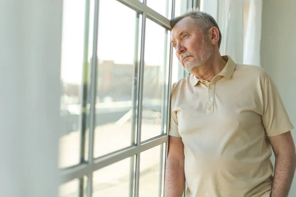 Stock image Sad tired lonely disappointed older senior man at home alone. Unhappy grandfather looking sadly outside window experiencing grief relative death bad news. Stressed elder man suffering from loneliness