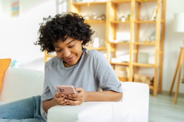 African girl holding smartphone touch screen typing scroll page at home. Woman with cell phone surfing internet using social media apps playing game. Shopping online Internet news cellphone addiction