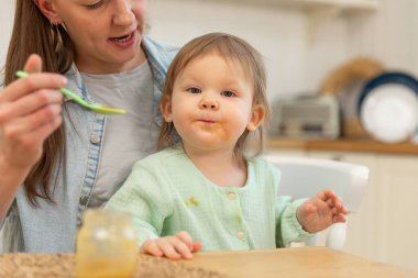 Evde mutlu bir aile. Anne mutfakta bebeğini kaşıkla besliyor. Dağınık suratlı küçük çocuk evde sağlıklı yiyecekler yiyor. Genç kadın anne, küçük kızına yemek veriyor.